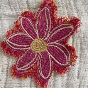 May include: A vibrant, flower-shaped coaster with a magenta center, outlined with cream-colored stitching. The petals have a textured appearance and are fringed with orange and yellow threads. The coaster is likely handmade.