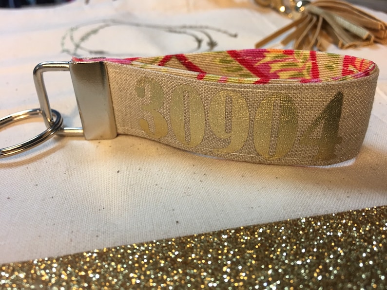 Op de afbeelding: Een stoffen polsbandje sleutelhanger met een gouden metallic nummerontwerp. Het polsbandje is beige met de nummers "30904" erop gedrukt. De binnenstof heeft een bloemenpatroon met roze, gele en groene kleuren. Een zilveren metalen sleutelring is bevestigd.