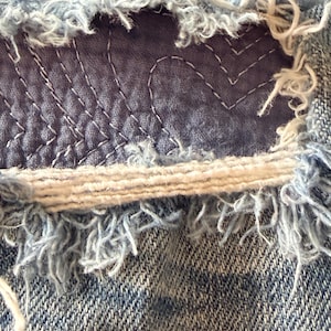 May include: A light blue denim fabric featuring a distressed, frayed horizontal opening. A dark blue fabric patch is visible underneath the tear, secured with white thread stitched in a repeating heart pattern across the surface of the patch.
