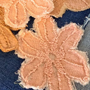 May include: Several light peach-colored fabric flower appliques with frayed edges and textured embroidery details. These floral shapes are layered over a dark blue denim fabric background, showcasing a contrast between the soft textile petals.