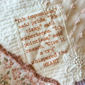 May include: A patchwork textile piece featuring cream, floral, and red-striped fabric segments. A central cream fabric square displays the printed text: "It's impossible," said pride. "It's risky," said experience. "It's pointless," said reason. "Give it a try," whispered the HEART.