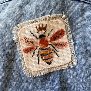 May include: A square, frayed-edge fabric patch featuring an embroidered bee with a crown, orange and black stripes, and pinkish-red wings. The patch is attached to a blue denim fabric. The embroidery includes small handwritten text near the wings.