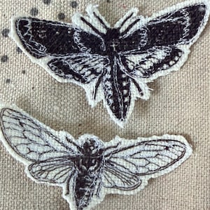 May include: Two embroidered fabric patches featuring insect designs. The top patch is a black moth with white wing details. The bottom patch is a black cicada with intricate white wing venation. Both patches have a white border on a beige fabric.