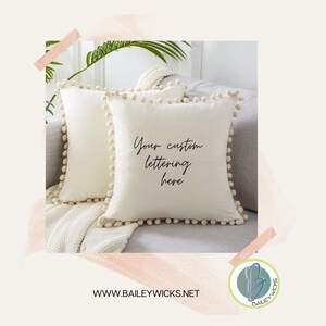 May include: Two cream-colored square throw pillows with a pom-pom trim. One pillow has the text "Your custom lettering here" in black script. The pillows are on a gray sofa with a white blanket.