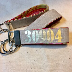 Op de afbeelding: Twee sleutelhangers met zilverkleurige hardware. E&eacute;n is lichtgrijs met het nummer "30904" in goud. De andere heeft een kleurrijk bloemenpatroon aan de binnenkant. Beide hebben een lus om gemakkelijk te dragen.