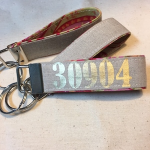 Op de afbeelding: Twee stoffen pols-sleutelhangers. De buitenkant is lichtgrijs met het nummer "30904" in goud gedrukt. De binnenstof heeft een roze, rode en gele bloemenprint. Elke sleutelhanger heeft een zilveren metalen sluiting en sleutelring.