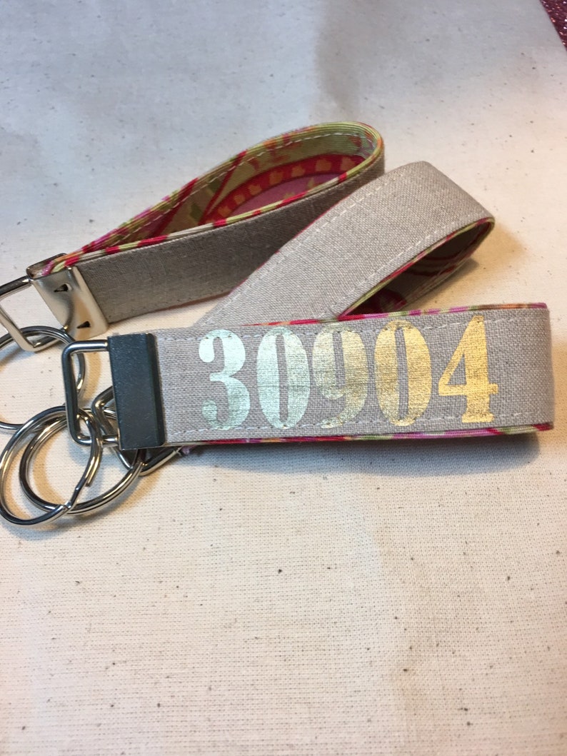 Op de afbeelding: Twee stoffen pols-sleutelhangers. De buitenkant is lichtgrijs met het nummer "30904" in goud gedrukt. De binnenstof heeft een roze, rode en gele bloemenprint. Elke sleutelhanger heeft een zilveren metalen sluiting en sleutelring.