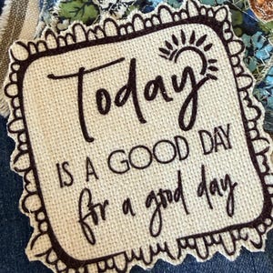 May include: A square fabric patch with a scalloped embroidered border in dark brown thread. The off-white canvas center features dark brown embroidered text that reads Today IS A GOOD DAY for a good day, accompanied by a small sun icon.