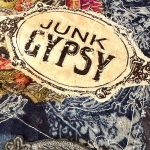 May include: A blue denim fabric surface featuring a central oval patch with the text JUNK GYPSY in black lettering. The textile is decorated with various embroidered floral and leaf motifs in shades of pink, green, and dark grey thread.