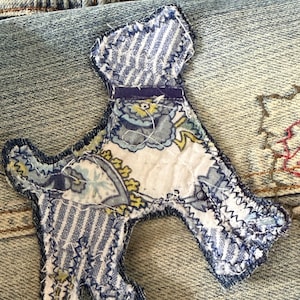 May include: A fabric applique in the shape of a dog, featuring a mix of blue and white striped fabric and a floral pattern with yellow and blue accents. The edges are finished with a dark blue zigzag stitch, and a thin dark blue ribbon collar.