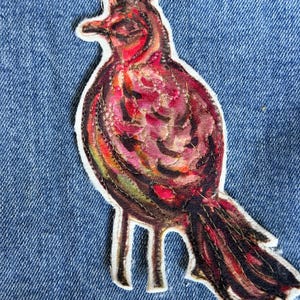 May include: A colorful bird patch with a white border, featuring shades of red, brown, and orange. The patch is sewn onto a blue denim fabric. The bird has a detailed design with visible stitching, creating a textured appearance. The patch is a decorative item.