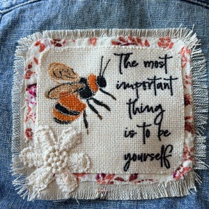 May include: A square fabric patch with frayed edges featuring an embroidered orange and black bee, a white crocheted flower, and the black embroidered text "The most important thing is to be yourself" on a cream-colored background.