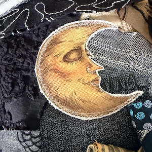 May include: A fabric patch featuring a golden crescent moon with a human face, stitched onto a background of various textured textiles including black fabric with white embroidery, grey patterned cloth, and striped beige fabric.
