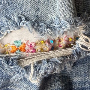 May include: Close-up of distressed denim fabric with a frayed edge. The denim features an embroidered floral design with orange, pink, and green thread, accented with colorful sequins. The fabric is light blue.