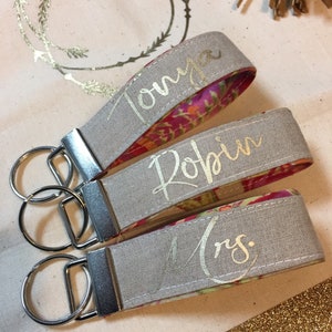May include: Three personalized keychains with silver hardware. The keychains have a gray exterior with gold lettering that reads "Tonya", "Robin", and "Mrs.". The interior fabric features a colorful floral pattern. The keychains are on a light beige surface.