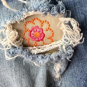 May include: Close-up of distressed denim jeans with a floral embroidery patch. The patch features a pink flower with an orange outline and center, stitched onto a tan fabric. The denim has frayed edges and a light blue wash.