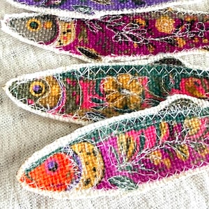 May include: Close-up of four colorful, embroidered fish. Each fish features a unique pattern with shades of purple, pink, green, and orange, outlined with white stitching. The fish are arranged on a textured, off-white fabric.