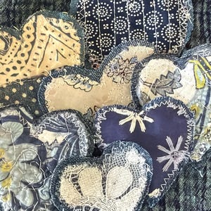 May include: A collection of heart-shaped fabric patches featuring various patterns including dark blue geometric prints, beige floral designs, and solid navy blue fabric. Each heart is outlined with visible white stitching and frayed denim edges.