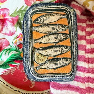 May include: A rectangular patch featuring a sardine can design. The patch has a denim blue border with white stitching. Inside, five fish are depicted on an orange background, with a lemon slice. The patch is set against a colorful floral and striped fabric.