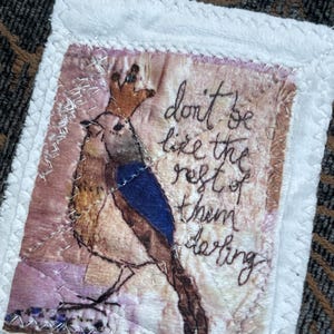 May include: A fabric patch with a bird illustration wearing a crown, featuring the text "don't be like the rest of them darling." The bird has blue and brown feathers. The patch is framed with white fabric and stitched with silver thread.