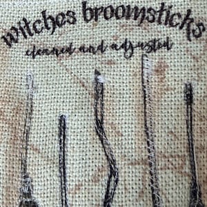 May include: A beige fabric patch featuring dark brown embroidered text that reads Witches broomsticks cleaned and adjusted. Below the text are five embroidered illustrations of broomsticks in varying shapes and sizes, rendered in dark brown thread.