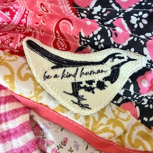 May include: A white patch with a black bird design and the words "be a kind human" stitched in black. The patch is set against a colorful patchwork background of pink, red, yellow, and floral patterns.