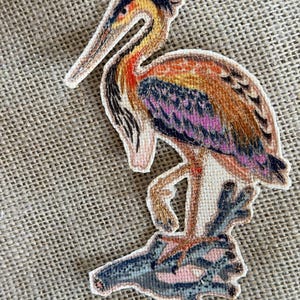 May include: A fabric patch featuring a detailed illustration of a heron standing on a dark, textured branch. The bird has a long, pointed beak, a yellow and orange neck, and wings with shades of brown, purple, and black. The patch has a die-cut edge.