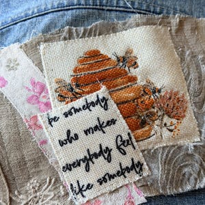 May include: A fabric patch featuring a printed beehive and bees on a beige background, layered over a piece of fabric with pink floral patterns. A smaller white fabric patch is stitched on top with the black embroidered text: Be somebody who makes everybody feel like somebody.