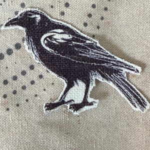 May include: A black and white fabric patch featuring a detailed illustration of a crow in profile. The bird is depicted with dark plumage and white highlights on its wing and chest, with a raw, frayed edge around the entire shape of the bird.