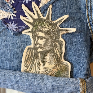 May include: Close-up of a denim garment with a patch featuring the Statue of Liberty. The garment is adorned with patches of red paisley, blue fabric with white star embroidery, and cream-colored lace. The denim has a rolled hem.