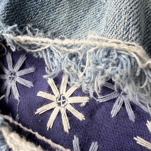 May include: Close-up of distressed denim fabric with frayed edges. A section of deep blue fabric is embroidered with white and light blue starburst designs. The denim is a light blue wash.