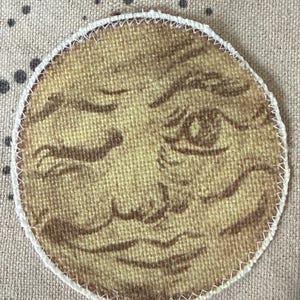 May include: A circular fabric patch featuring a tan and brown illustration of a human-like moon face with closed eyes and a smiling mouth. The patch has a visible woven texture and is finished with a white zigzag stitched border around the edge.