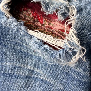 May include: Close-up of distressed blue denim jeans with a large tear revealing a patterned fabric underneath. The edges of the tear are frayed, with white and blue threads. The fabric visible through the tear has a floral design.