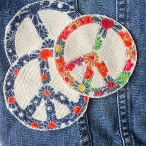 May include: Three embroidered peace sign patches in a circular shape. The patches are made of a natural-colored fabric with colorful floral designs. The patches are placed on a blue denim jacket.
