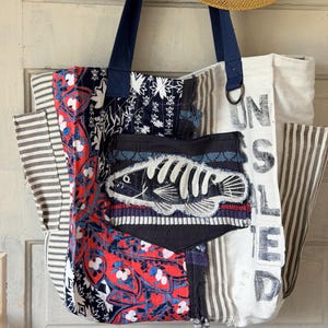 May include: A patchwork tote bag with navy blue handles and a fish skeleton applique. The bag features various fabric patterns, including floral, striped, and a white fabric with the word "UNSALTED" printed on it. A straw hat hangs above the bag.