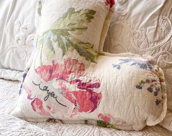 May include: A cream-colored, heart-shaped pillow with a floral pattern in pink, red, and green. The pillow has the words "i love you" printed on a fabric strip. The pillow is placed on a white, quilted bedspread.