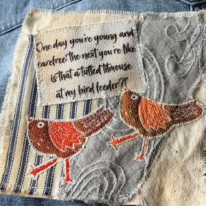 May include: A fabric patch featuring two embroidered birds with orange and brown wings and red legs. The patch includes a rectangular piece of off-white fabric with the black text: One day you're young and carefree. The next you're like is that a tufted titmouse at my bird feeder?!