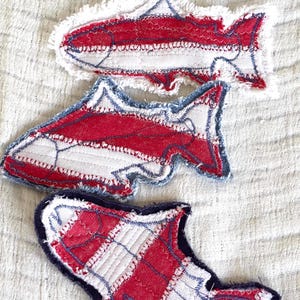 May include: Three handmade fabric fish appliqués. Each fish features a red and white striped pattern with blue stitching and a frayed edge. The fish are arranged on a textured, off-white background.