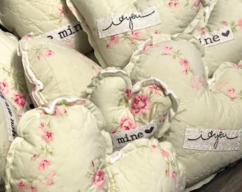 May include: A collection of heart-shaped, quilted fabric ornaments. The ornaments are a pale green with a pink floral pattern. Each heart has a white ribbon with the words "I you" or "mine" in black script.