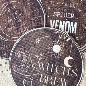May include: Three circular coasters with a dark brown, textured background. One features a witch hat, a cat, a sun, a moon, and the text "WITCH'S BREW CRAFTING SINCE 1782". Another displays "EYE OF NEWT". The third shows "PREMIUM QUALITY SPIDER VENOM".