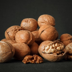 Puede incluir: Un montón de nueces marrones, algunas con las cáscaras abiertas, revelando los granos blancos en el interior. Las nueces están dispuestas en forma de pirámide sobre un fondo oscuro.
