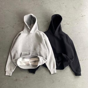 May include: Two hooded sweatshirts, one gray and one black, are laid out on a gray concrete surface. Both sweatshirts have a pouch pocket on the front.