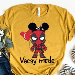 May include: Mustard yellow t-shirt featuring a cartoon Deadpool character with Mickey Mouse ears holding red balloons. The text "Vacay mode" is printed below the character. The shirt is paired with blue jeans and striped flats.