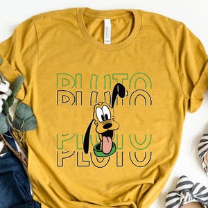 Könnte beinhalten: Ein senfgelbes T-Shirt mit einem Cartoon-Bild von Pluto dem Hund. Der Text "Pluto" wird in grünen Buchstaben auf dem Shirt wiederholt.
