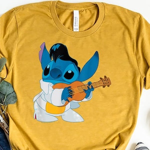 May include: Mustard yellow t-shirt featuring a blue cartoon character playing a ukulele. The character is dressed in a white jumpsuit and has black hair. The shirt is styled with blue jeans, striped flats, and decorative pumpkins.