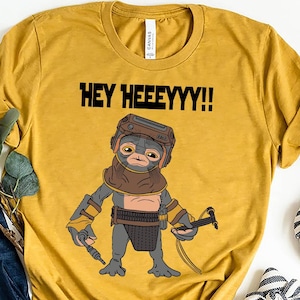 May include: Mustard yellow t-shirt with a cartoon alien character and the text "HEY HEEEEYYY!!" in black. The shirt is styled with blue jeans, striped flats, and decorative pumpkins. The shirt is made of a soft, comfortable material.