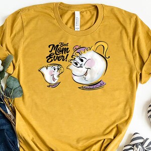 Può includere: T-shirt giallo senape con un'illustrazione a cartoni animati di Mrs. Potts e Chicco de La Bella e la Bestia, con la scritta "Best Mom Ever!". La maglietta è abbinata a jeans blu e ballerine a righe.