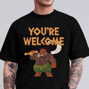 May include: Black t-shirt featuring the phrase "YOU'RE WELCOME" in orange text. The shirt displays a cartoon illustration of a Polynesian figure holding a weapon. The figure is brown with tattoos and a green grass skirt.
