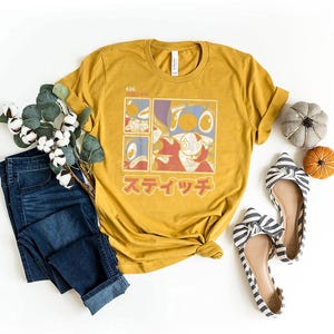 May include: Mustard yellow t-shirt with a cartoon character graphic in a grid design and Japanese text. The image includes blue jeans, striped flats, and decorative pumpkins.