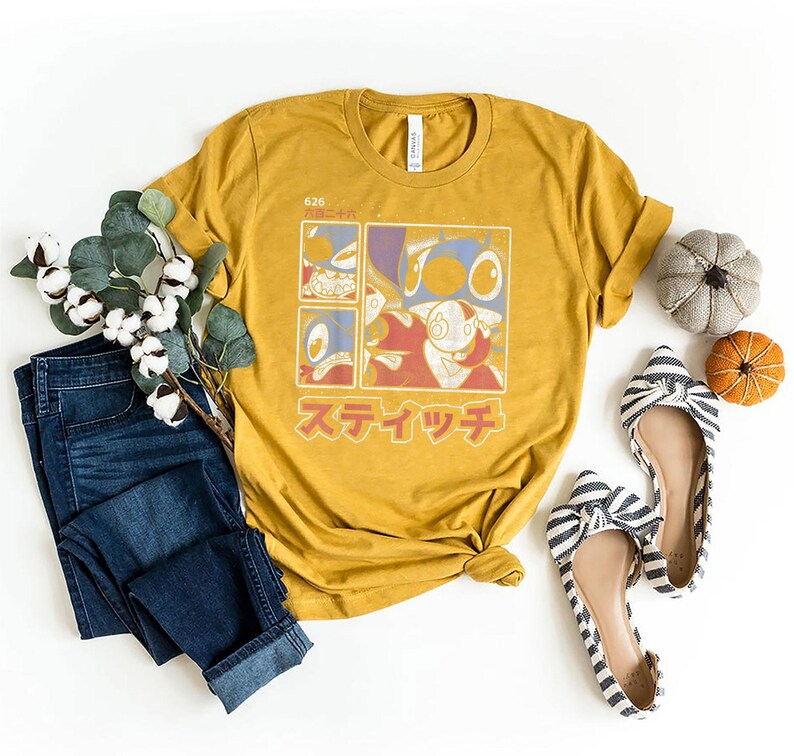 May include: Mustard yellow t-shirt with a cartoon character graphic in a grid design and Japanese text. The image includes blue jeans, striped flats, and decorative pumpkins.